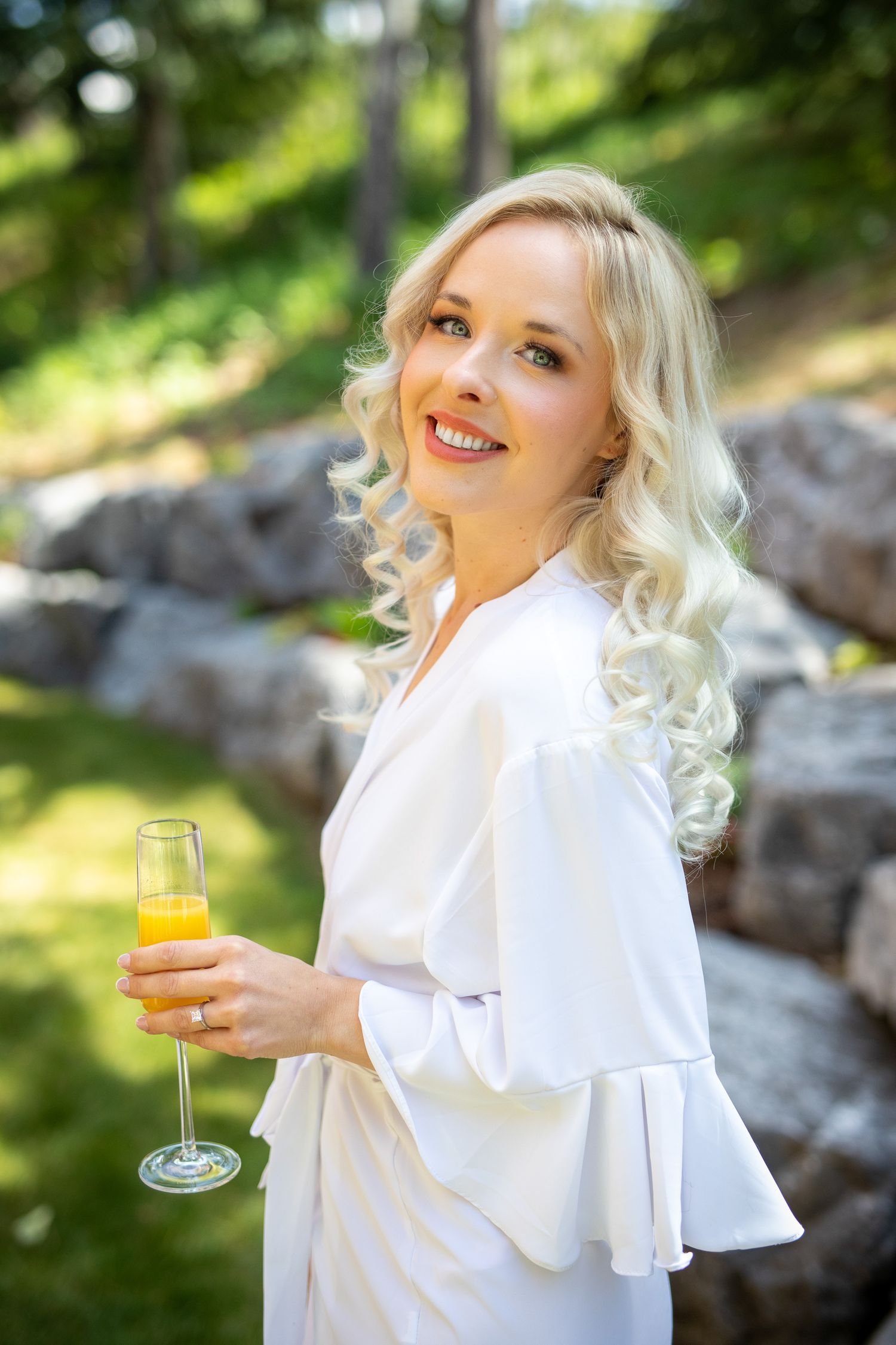 Smiling blonde woman in white dress holding a champagne flute with orange juice outdoors In Canmore