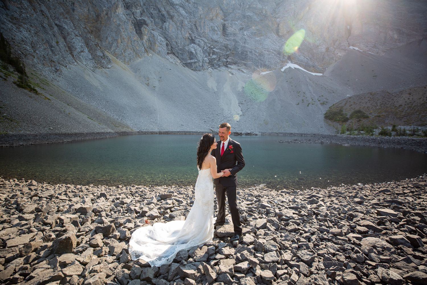 Bride and groom share a romantic moment on rocky shore beside a mountain lake with dramatic cliffs and sunlightin Kananaskis, Alberta.
