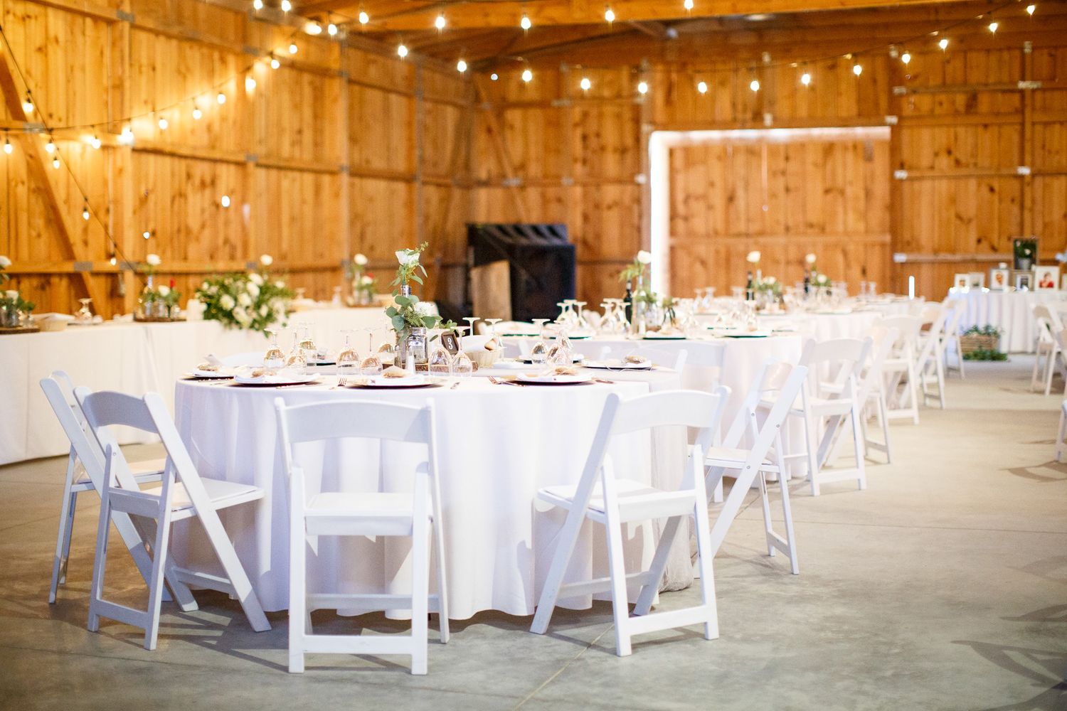 Elegant barn wedding reception with white round tables, folding chairs, floral centerpiece, and string lights overhead. Cherry creek estate