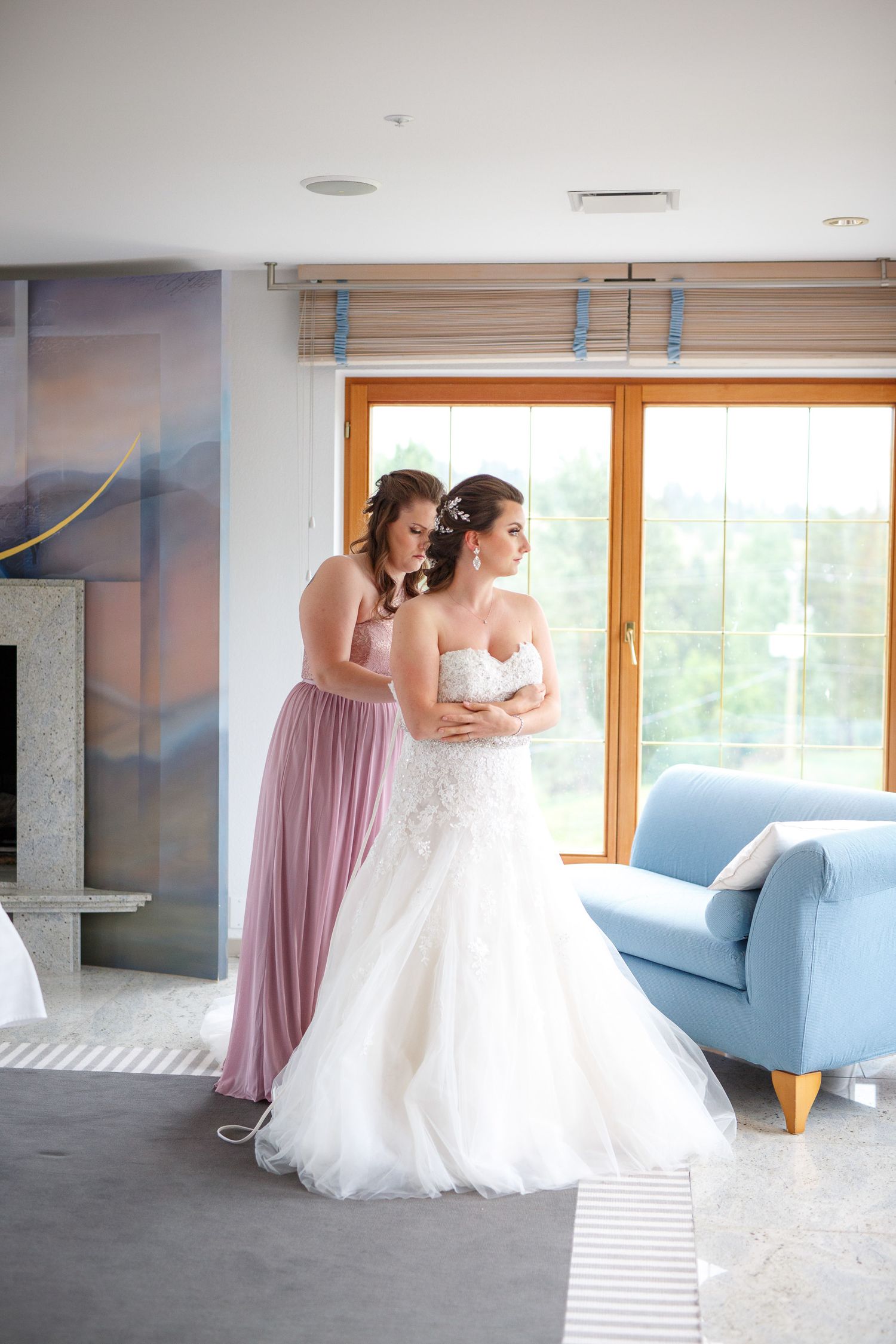 Bridesmaid in pink dress helping bride in white lace gown get ready in bright hotel room before wedding ceremony.
