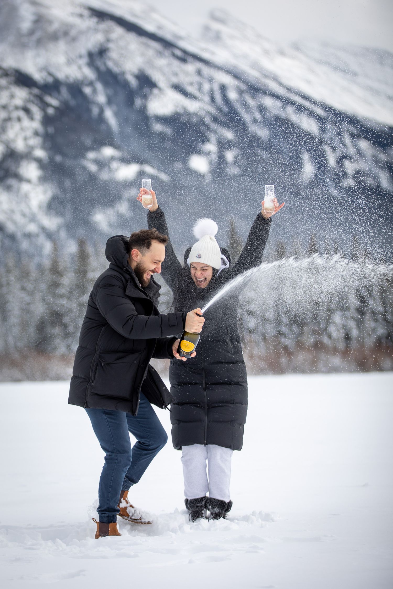A couple newly engaged celebrates with champagne in the snow against a backdrop of the majestic Mount Rundle, Vermilion Lakes Banff, Alberta
