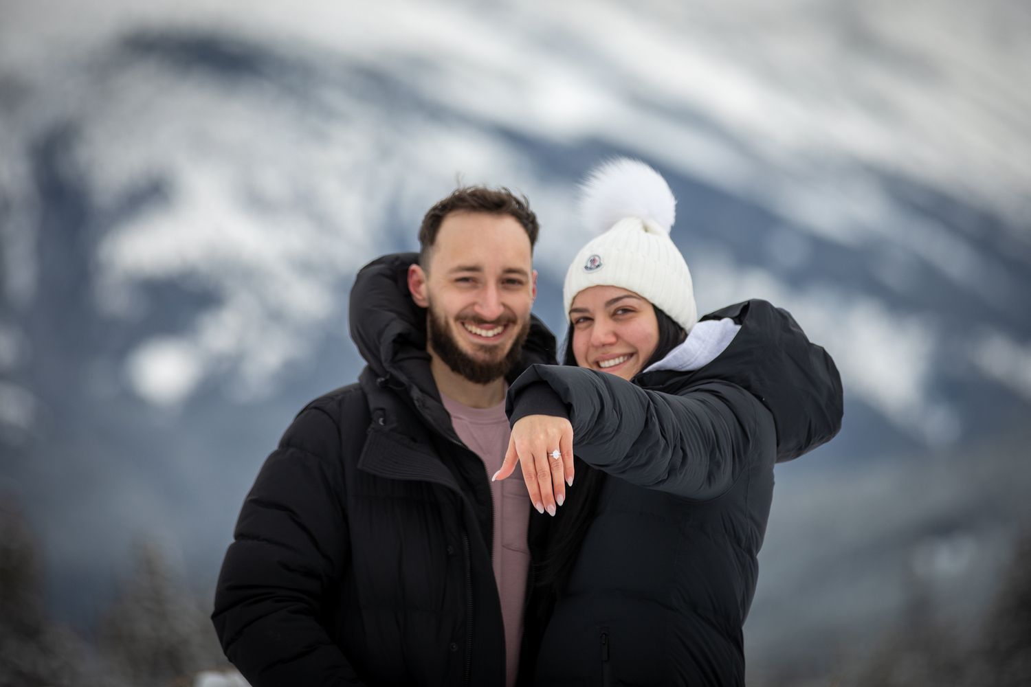 A couple embraces while smiling in front of a snowy mountain landscape on a cloudy winter day. Vermilion lakes in Banff. AB