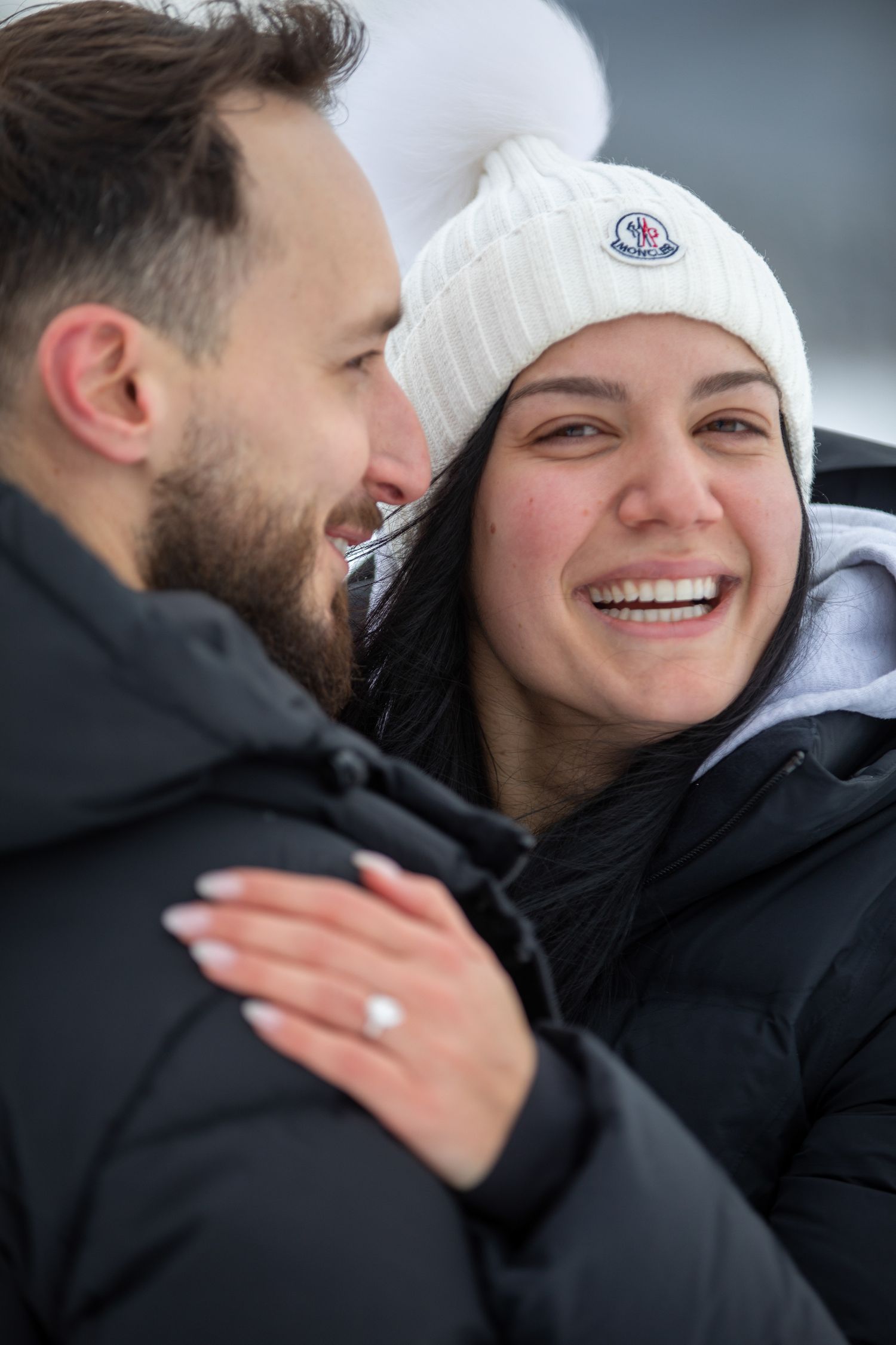 A couple in black winter coats share a joyful moment outdoors, with one wearing a white Moncler beanie.  Vermilion lakes in Banff. AB