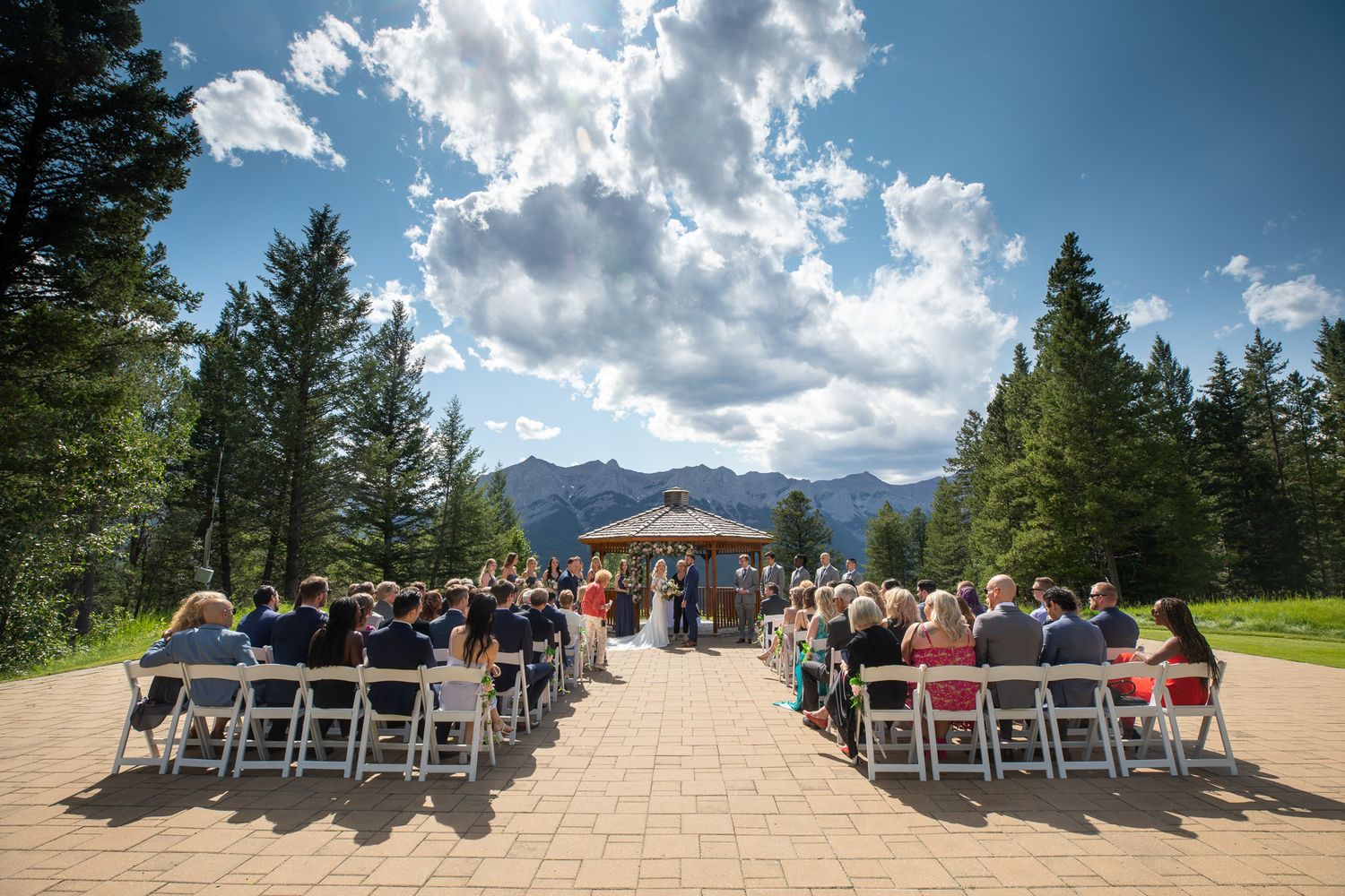 Outdoor mountain wedding ceremony with guests seated on white chairs facing a gazebo, surrounded by pine trees and scenic peaks at Silvertip