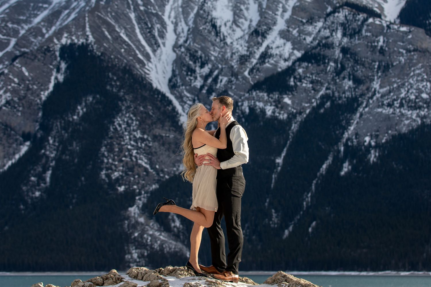 A couple embraces on a mountain peak with snow-capped mountains and dramatic winter landscape in the background at Lake Minnewanka in Banff.