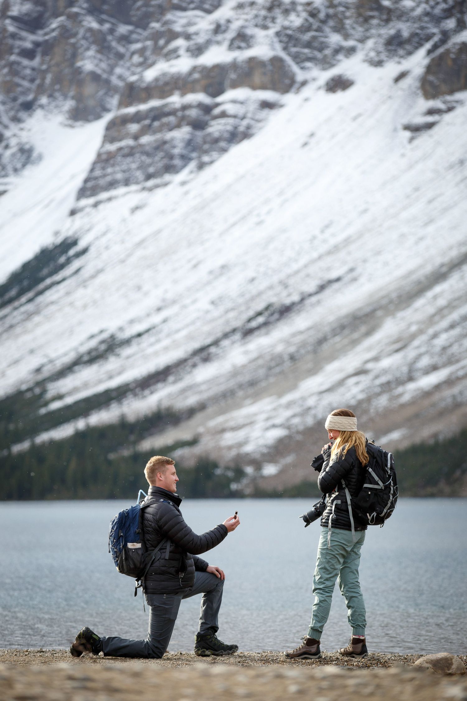 Surprise proposal on a rocky shore overlooking a scenic Bow lake in Banff National Park surrounded by snow-capped peaks in winter.