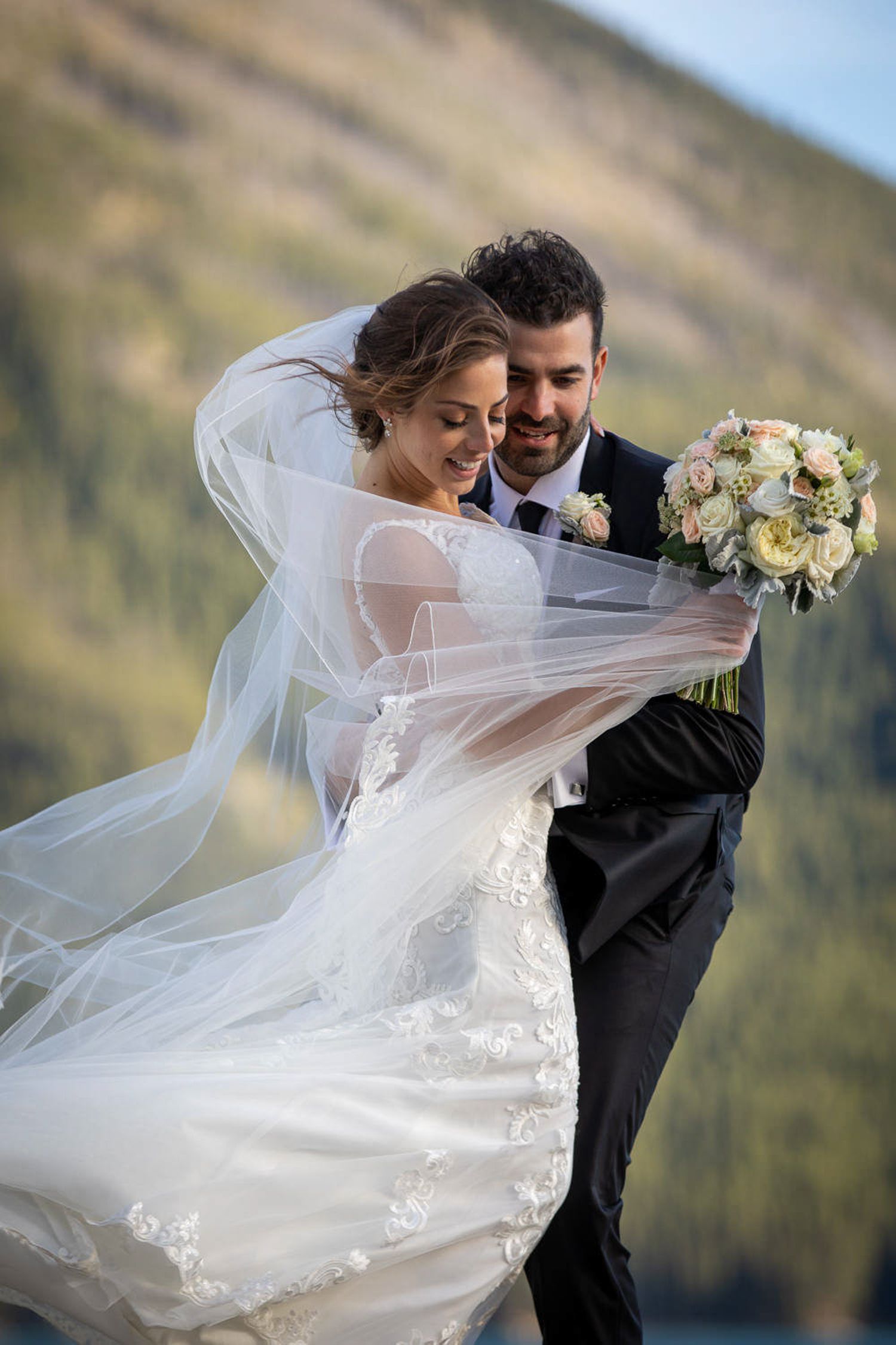 A bride and groom dance together outdoors with flowing white veil and mountain scenery in the background. Banff