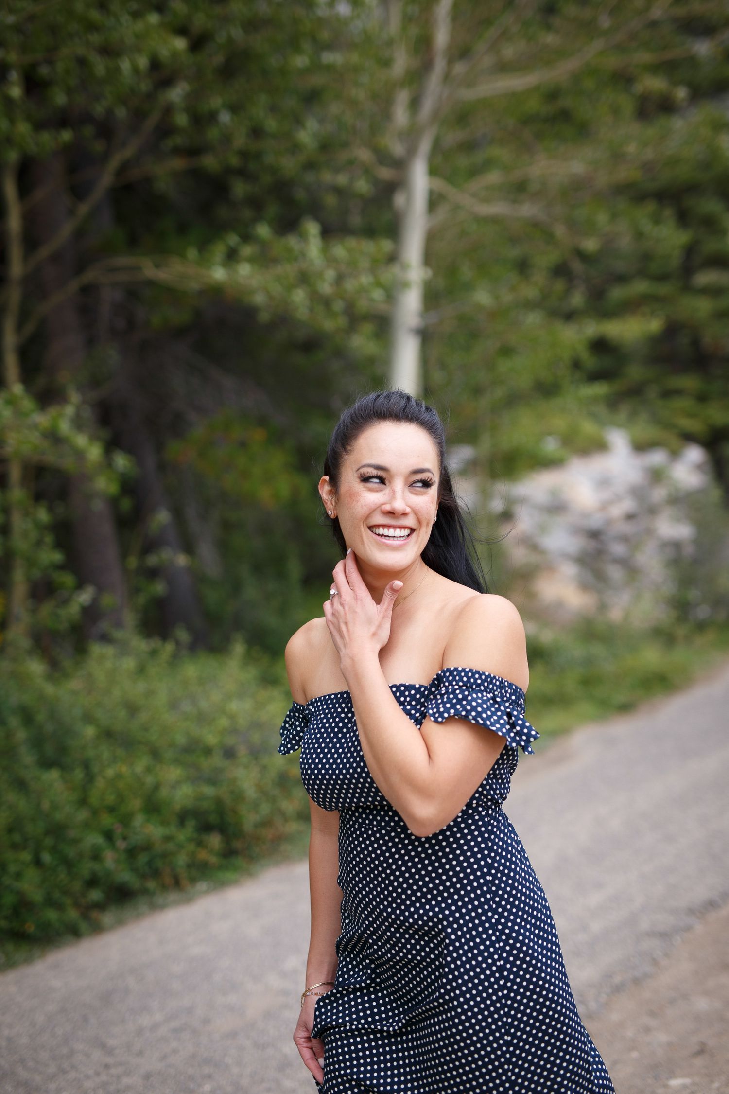 A smiling woman wearing a polka dot dress stands on a path surrounded by trees and greenery at Lake Minnewanka in Banff.