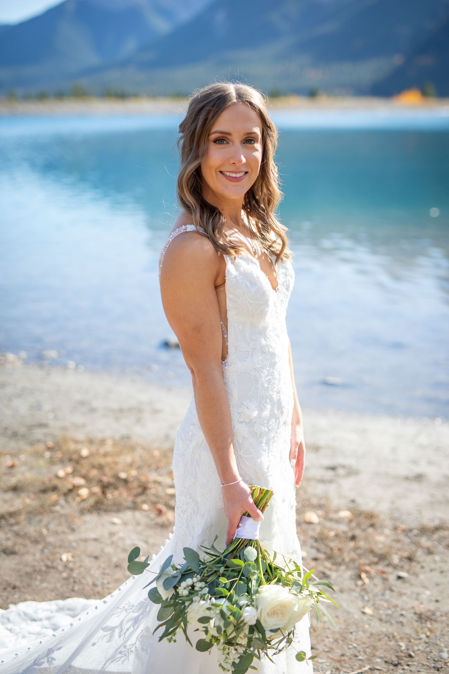 A bride in a white lace wedding dress holds a green and white bouquet while posing by a scenic mountain lake at Quarry lake in Canmore, AB.