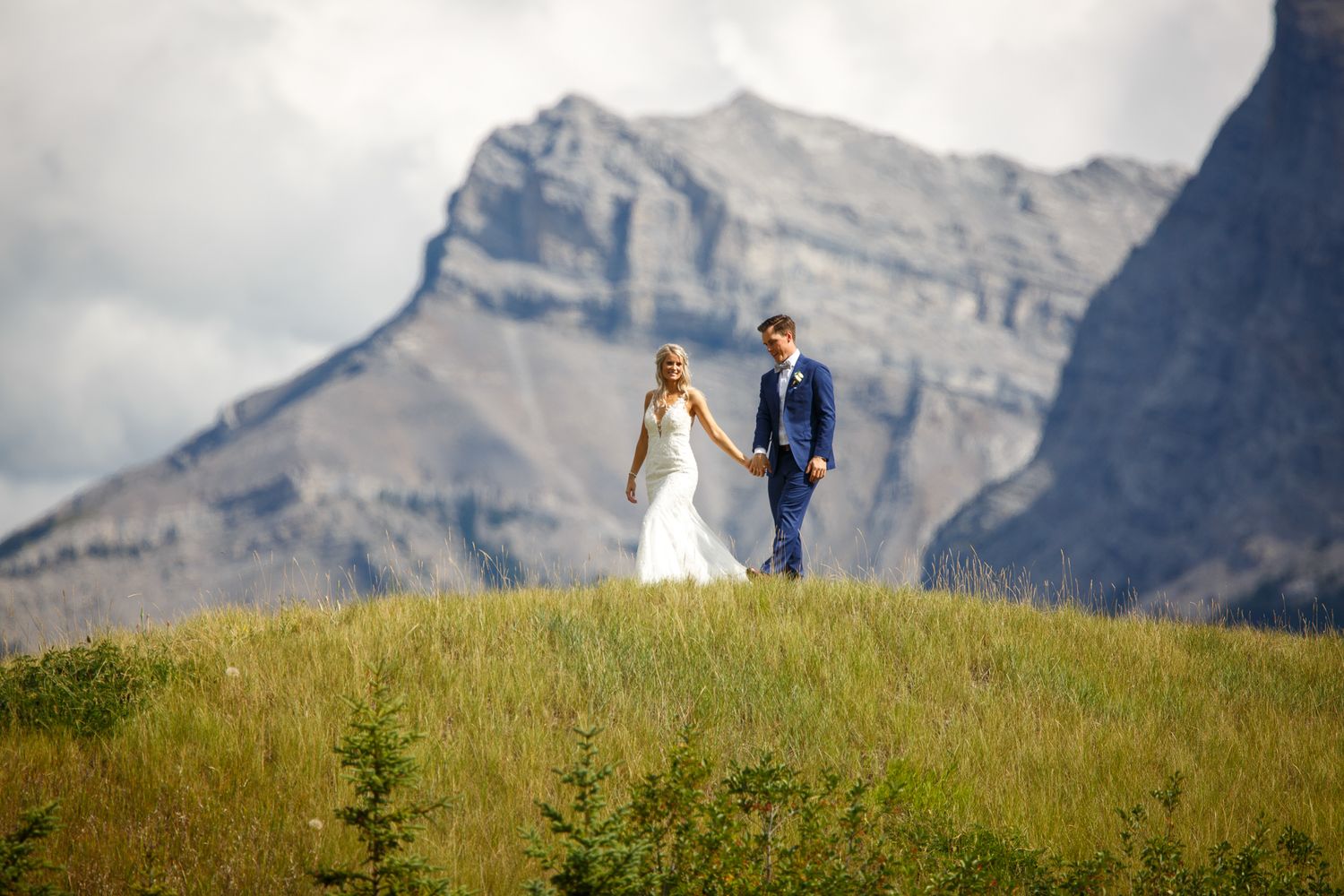 A couple in wedding attire stands on a grassy hillside against dramatic mountain peaks in Banff National Park.