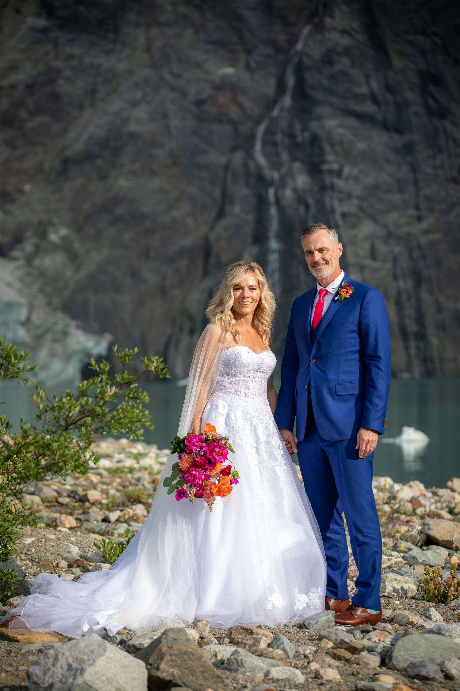 Bride in white gown and groom in blue suit pose by glacial lake with waterfall backdrop on rocky shore. Copperpoint resort,  Invermere.