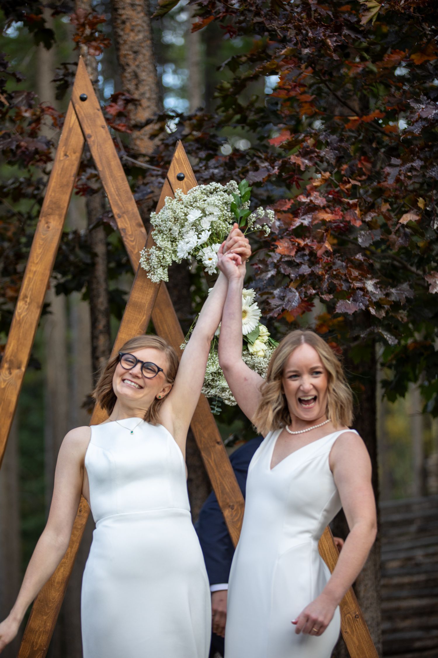 A LGBTQ couple joyful outdoor wedding scene with two people in white dresses celebrating under wooden arches with delicate flowers.