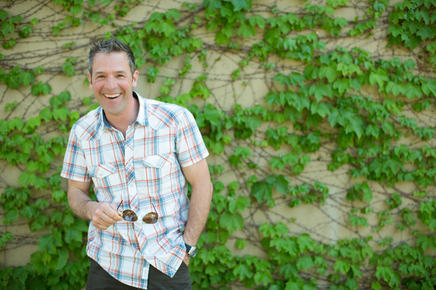 A smiling man in a plaid shirt stands in front of a green leafy background in Canmore, Alberta.