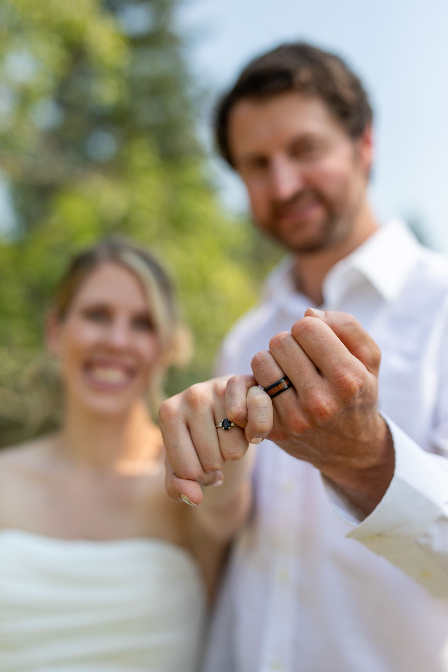 Smiling newlywed couple holding hands and showing off their wedding rings outdoors with green trees in background. Invermere, BC