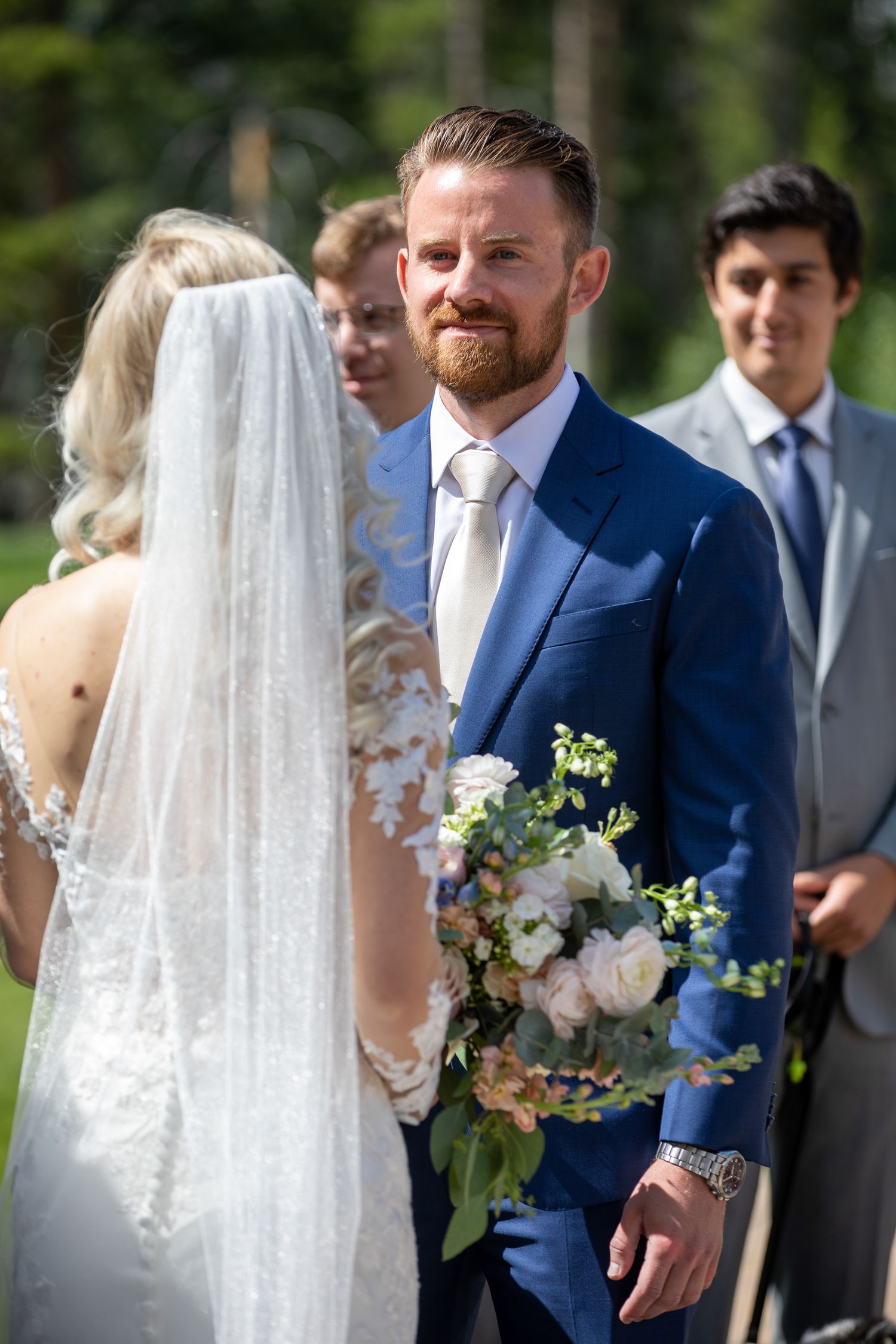 Groom in blue suit smiling at bride during outdoor wedding ceremony, holding bouquet of white and pink roses In Canmore Silvertip gazebo.