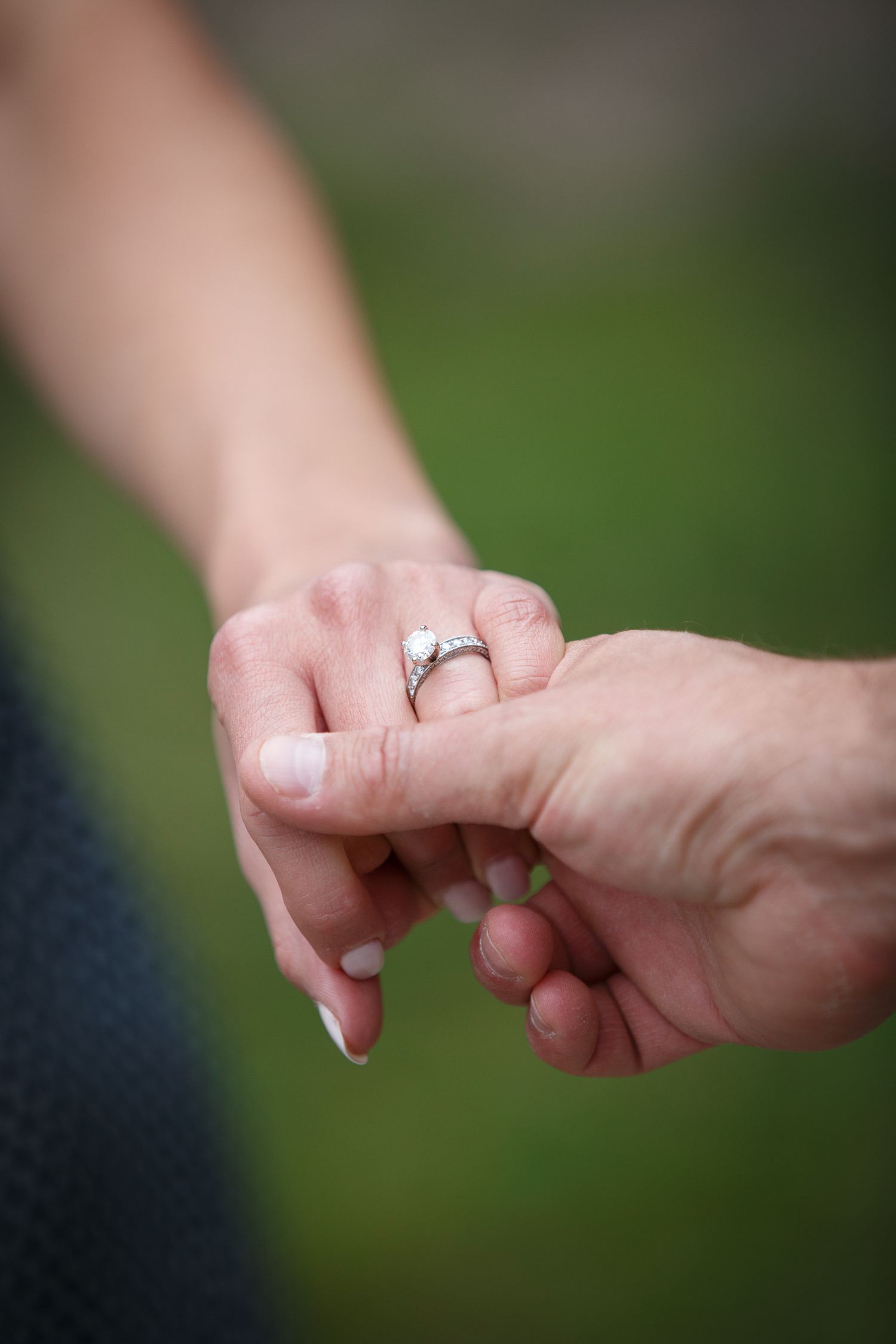 Two hands clasped together, with a ring visible on one of the fingers, against a blurred green background.