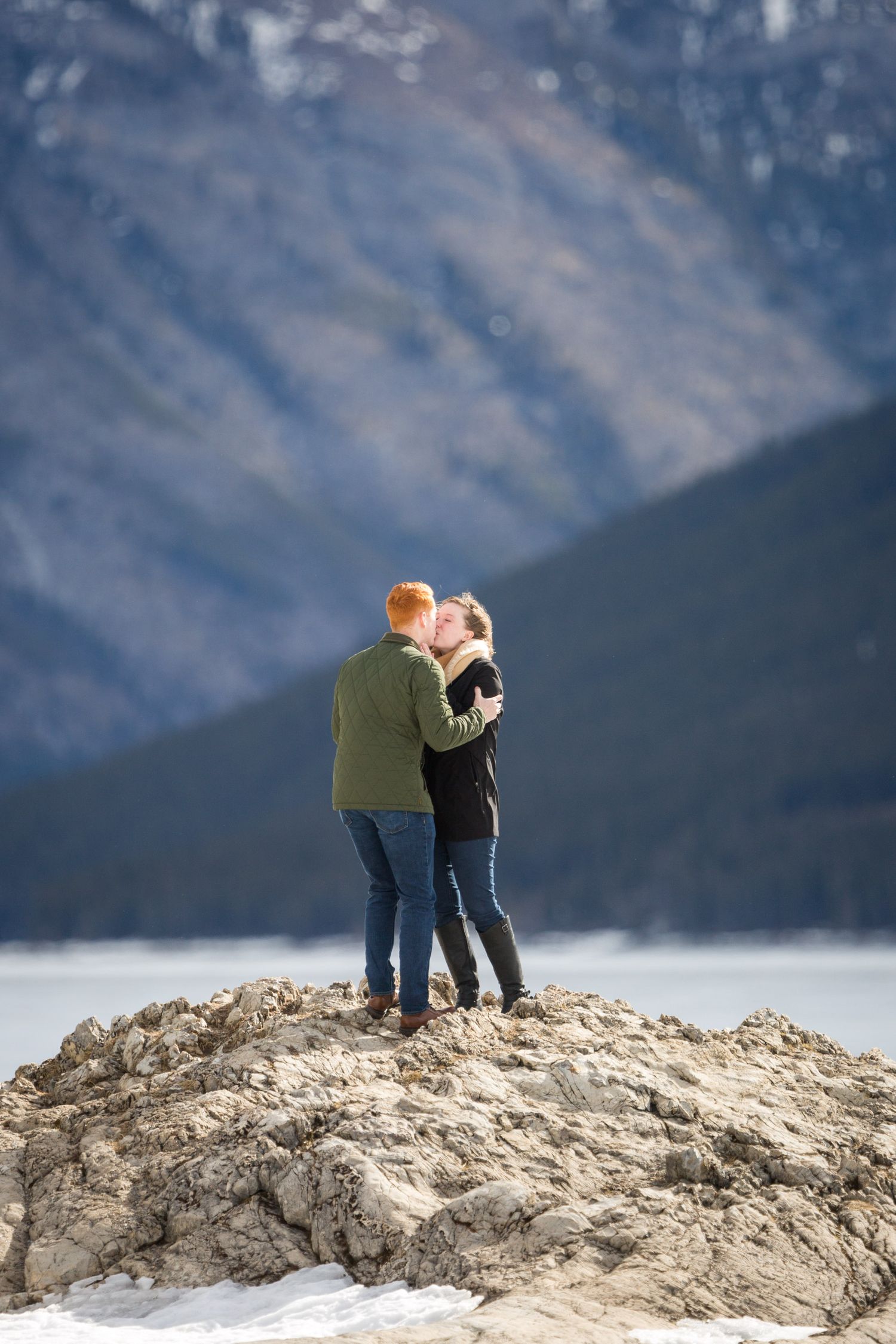 Couple kissing on rocky outcrop with snow-dusted mountains and frozen lake in background at Lake Minnewanka in Banff National Park.