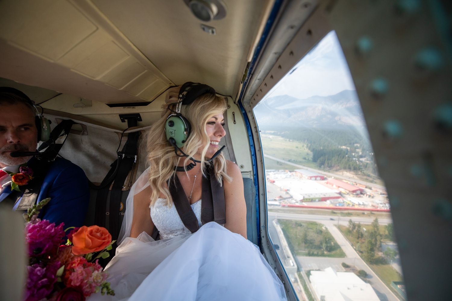 Bride in white dress wearing headphones smiles out helicopter window holding colourful bouquet on wedding day. Invermere, BC.