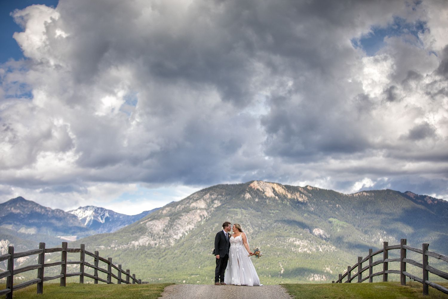 A bride and groom stands between wooden fences with dramatic mountain peaks in the background. Brady Creek ranch. Invermere, BC.