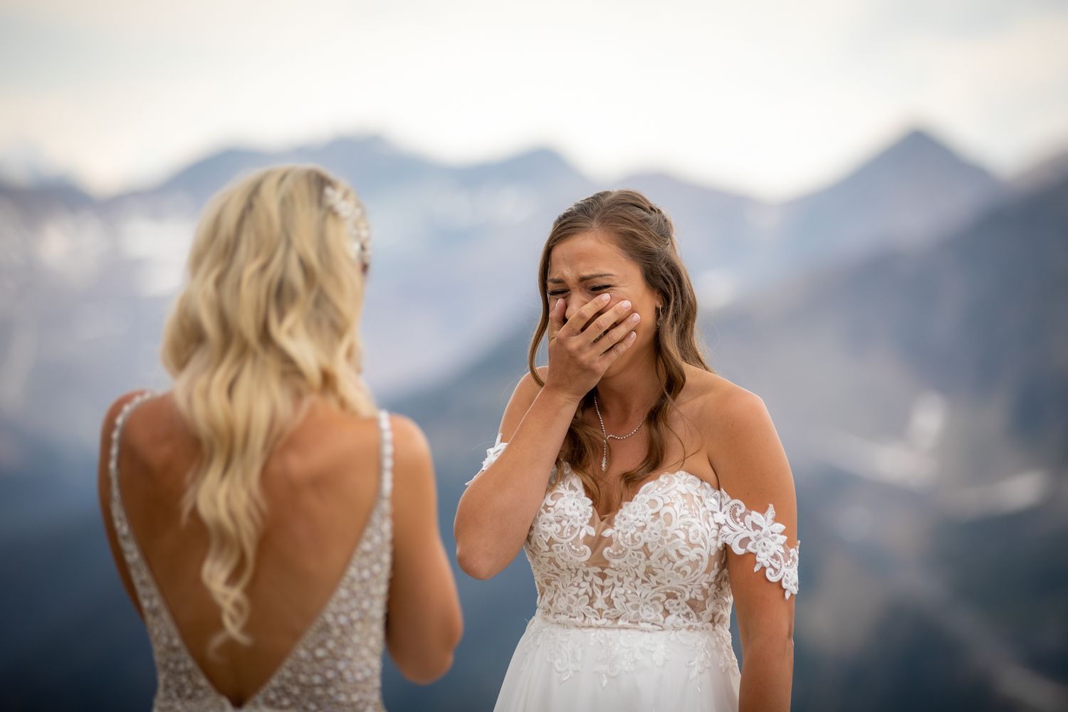 Two brides in lace wedding gowns share an emotional moment with mountain peaks in the background at Kicking Horse Resort, Golden, BC