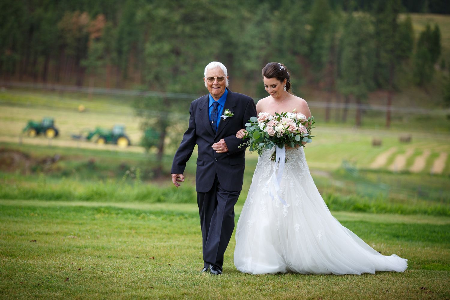 Bride in white gown walks with older gentleman in navy suit across green lawn with trees and tractors in background Cherry creek estate