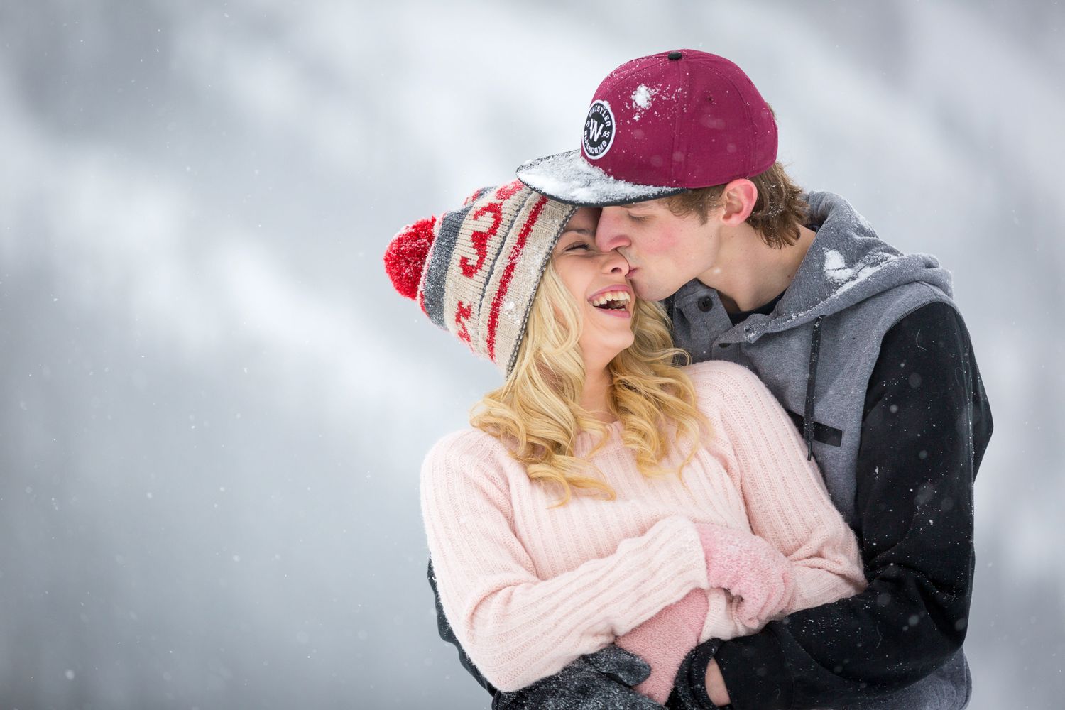 A couple shares a romantic moment in the snow, with her in pink and him wearing a red baseball cap at Lake Louise, AB