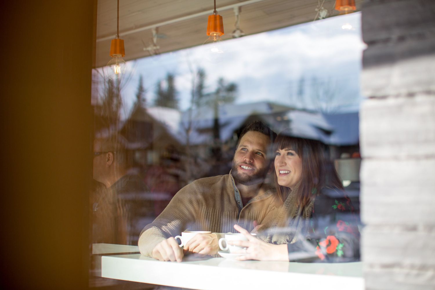 A couple sits together at a cafe table by a window, smiling while having coffee at Whitebark cafe in Banff, AB.