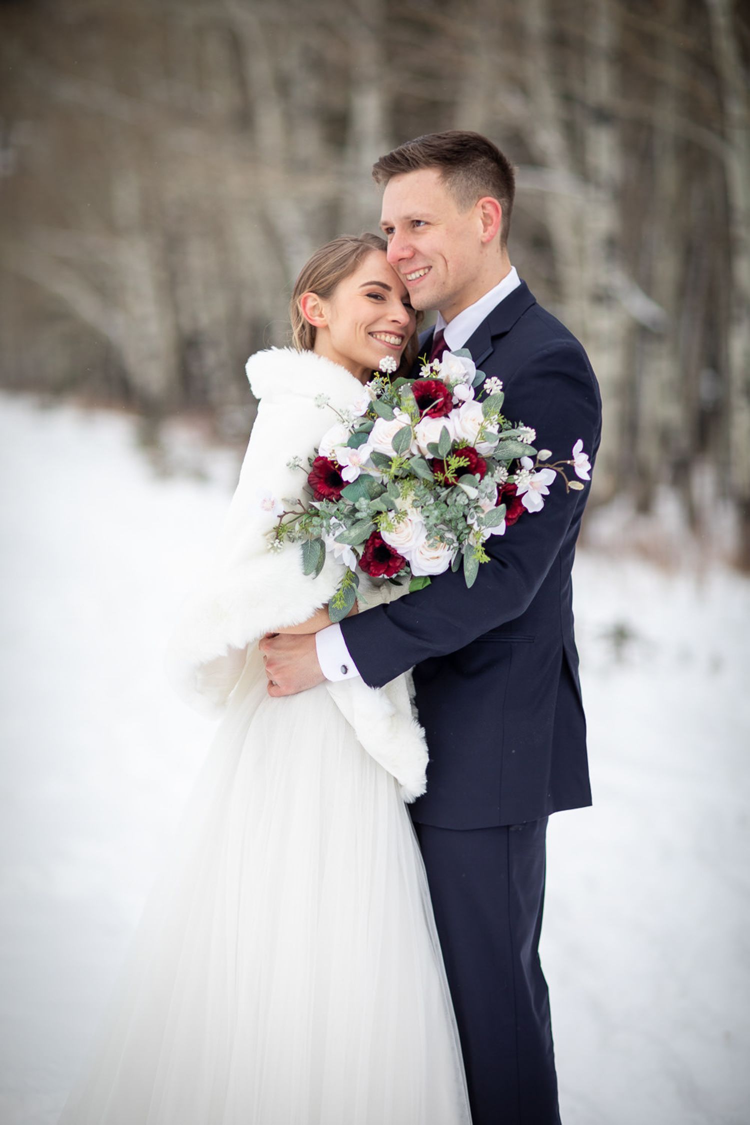 A romantic winter wedding photoshoot with a couple in navy suit and white dress holding a burgundy and white bouquet Benchland trail Canmore