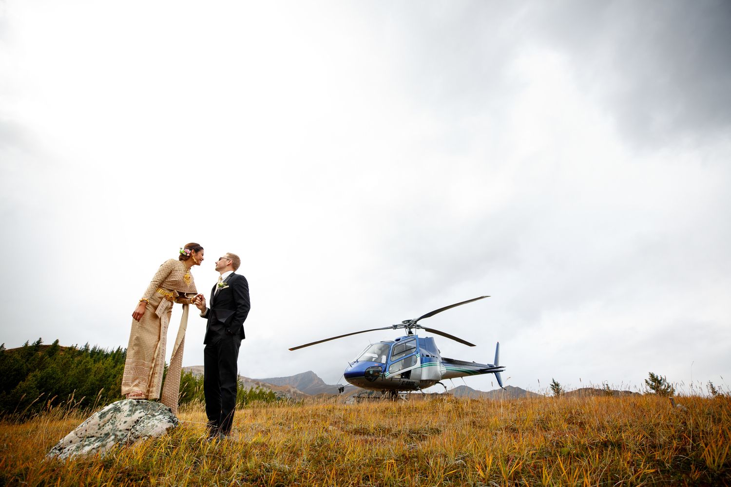 A wedding couple stands on a hilltop near a parked helicopter during a dramatic cloudy sunset. Cline river, AB