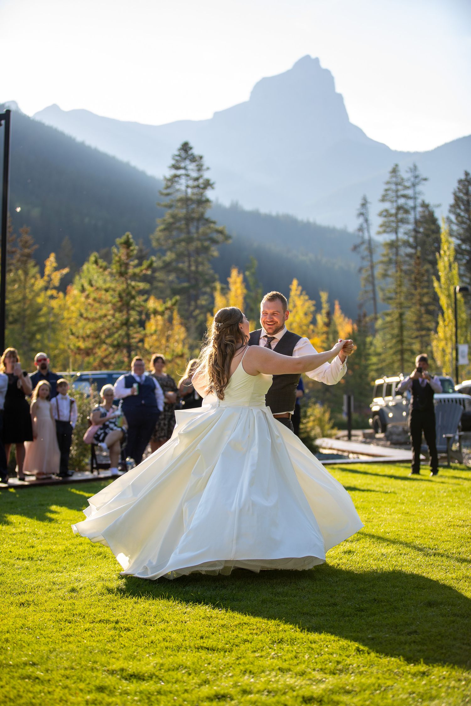 A couple dances on a green lawn with majestic mountain peaks and golden autumn trees in the background. Stewart Creek resort, Canmore, AB.