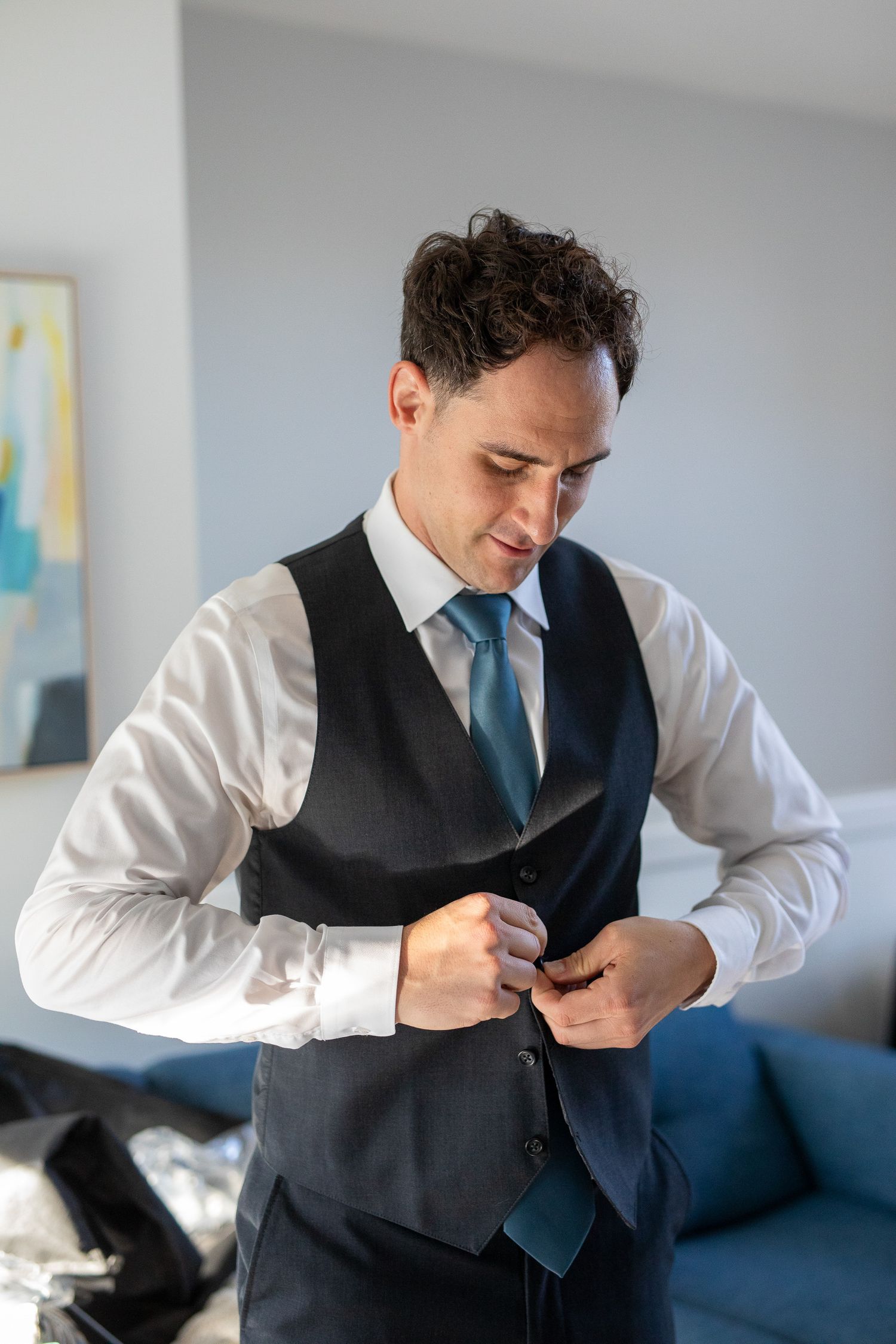 A groom in a white shirt and grey vest buttons up his formal attire while getting dressed.