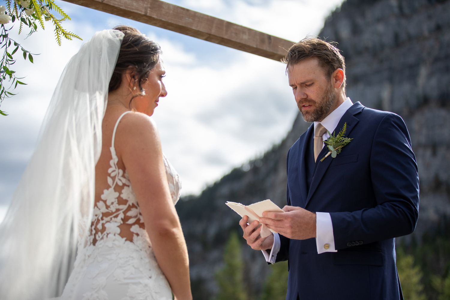 A couple exchanging vows during an outdoor mountain wedding ceremony beneath a wooden arch. Tunnel Mountain Reservoir Banff, AB