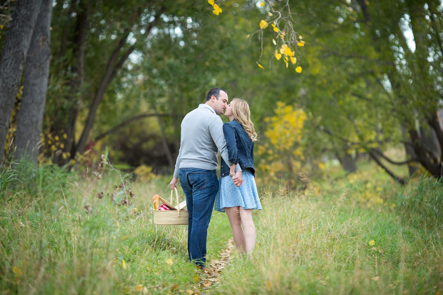 A couple embraces in a sunlit meadow surrounded by tall grass and autumn trees in Fish Park, Calgary, AB.