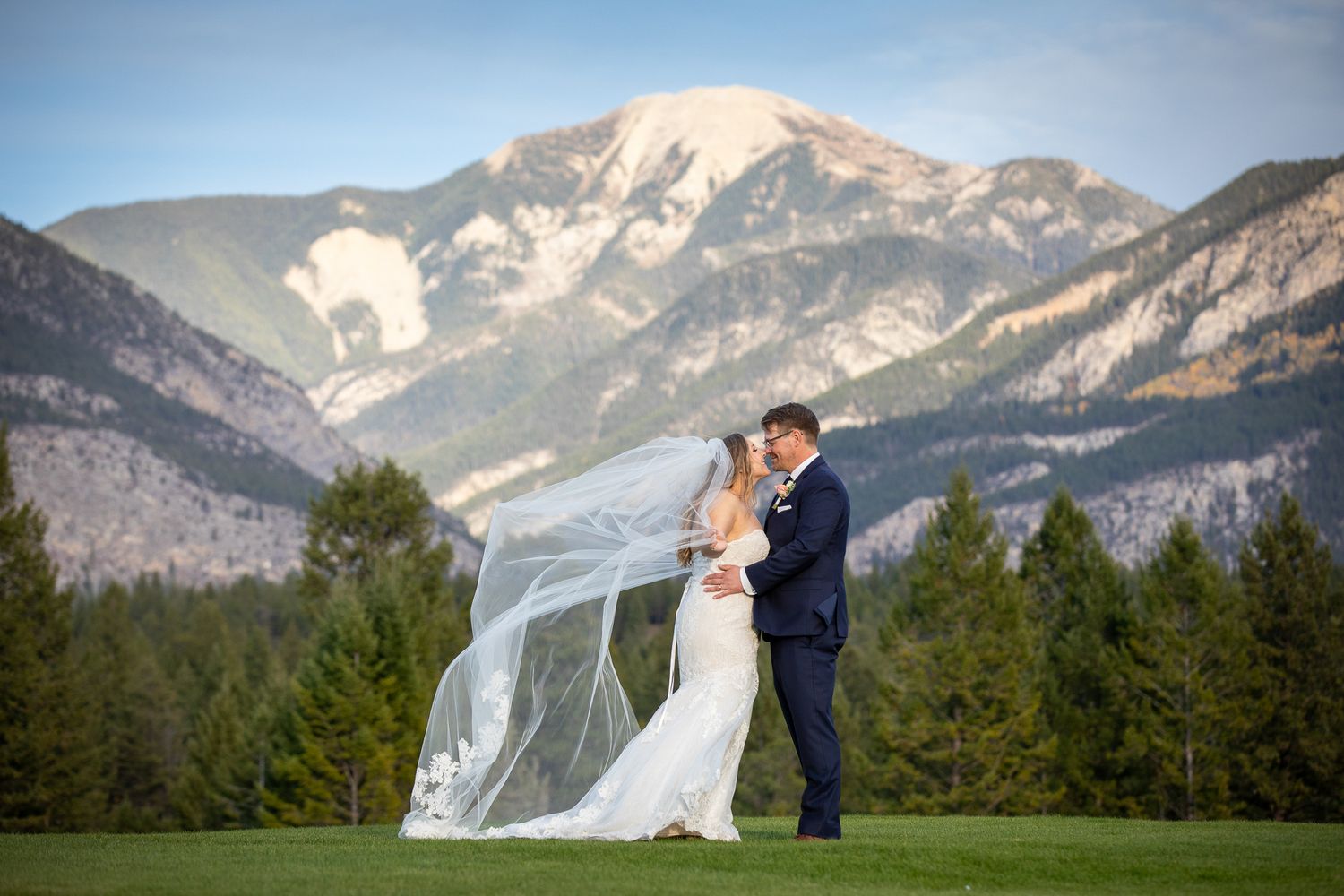 A couple shares a romantic kiss against the stunning backdrop of snow-capped mountain peaks and forests. Eagle Ranch Resort, Invermere, BC.