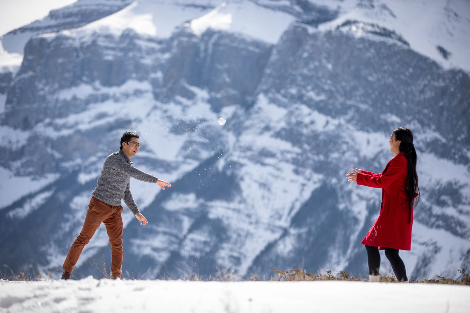 Two people playfully throw snowballs at each other against a backdrop of snow-covered mountain peaks in Banff, AB.