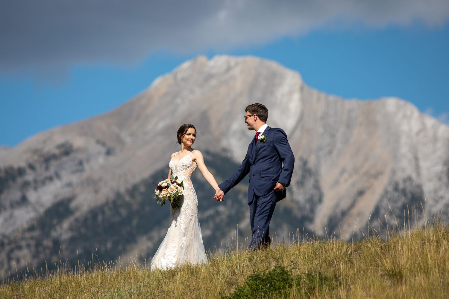 A couple in wedding attire stands together against a backdrop of majestic mountain peaks on a grassy hillside in Canmore Three Sisters area.