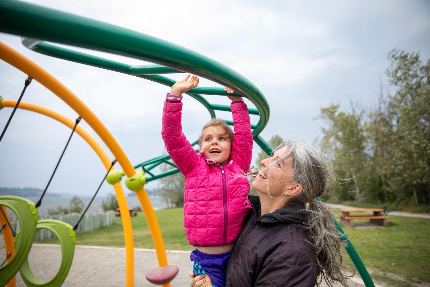 A person in a pink jacket plays on a curved yellow and green playground structure in a park setting Chabot Park in Invermere, BC.