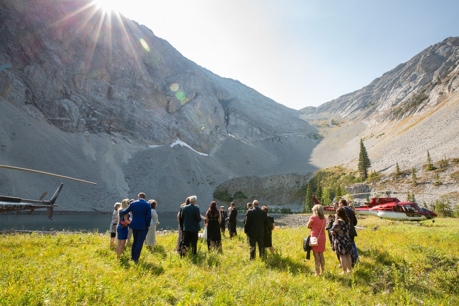 Group of people gathered in alpine meadow with helicopters and dramatic mountain backdrop on sunny day in Kananaskis, Alberta.