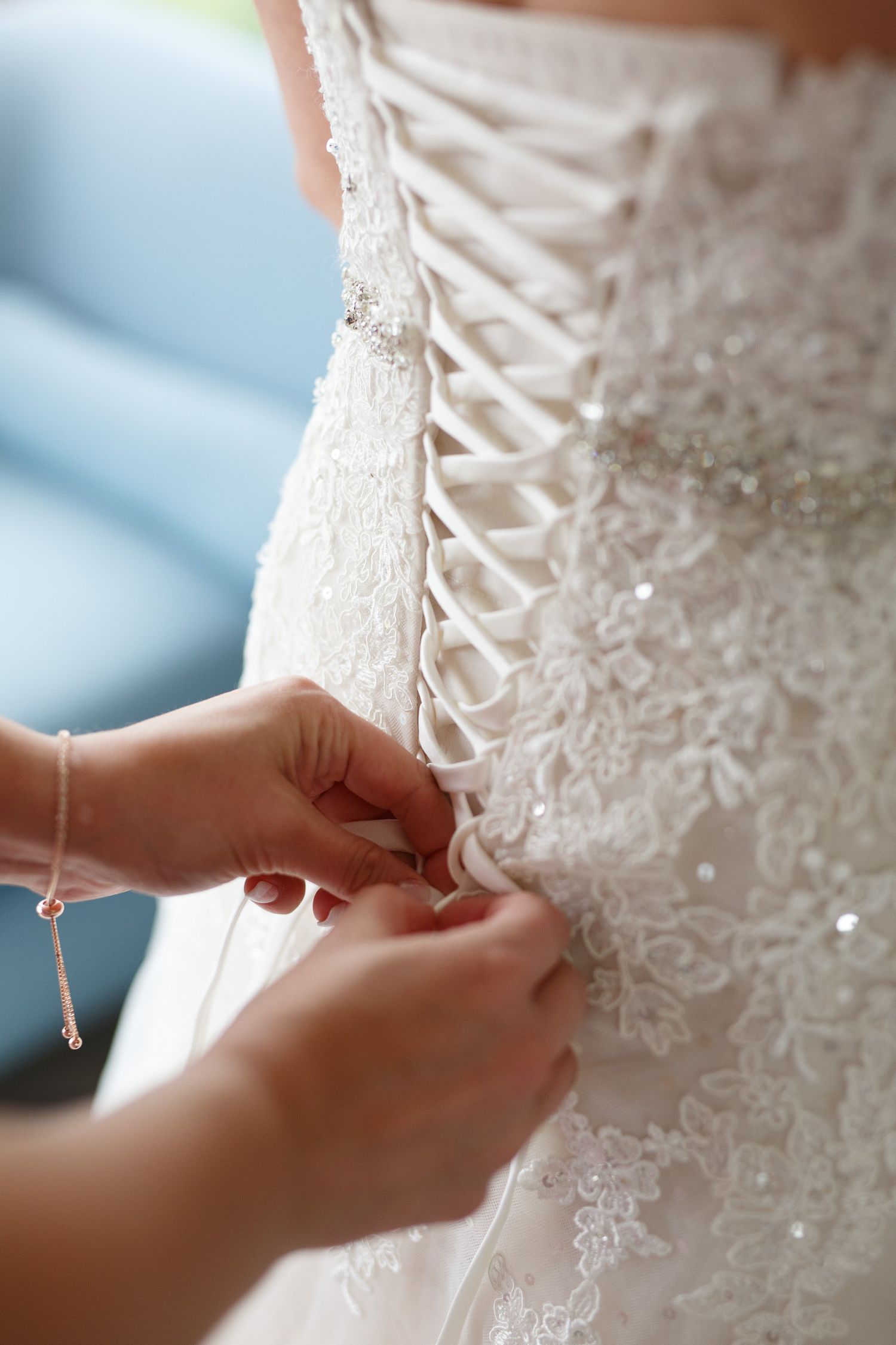 Hands lacing up a white beaded corset wedding dress, close-up bridal preparation detail.