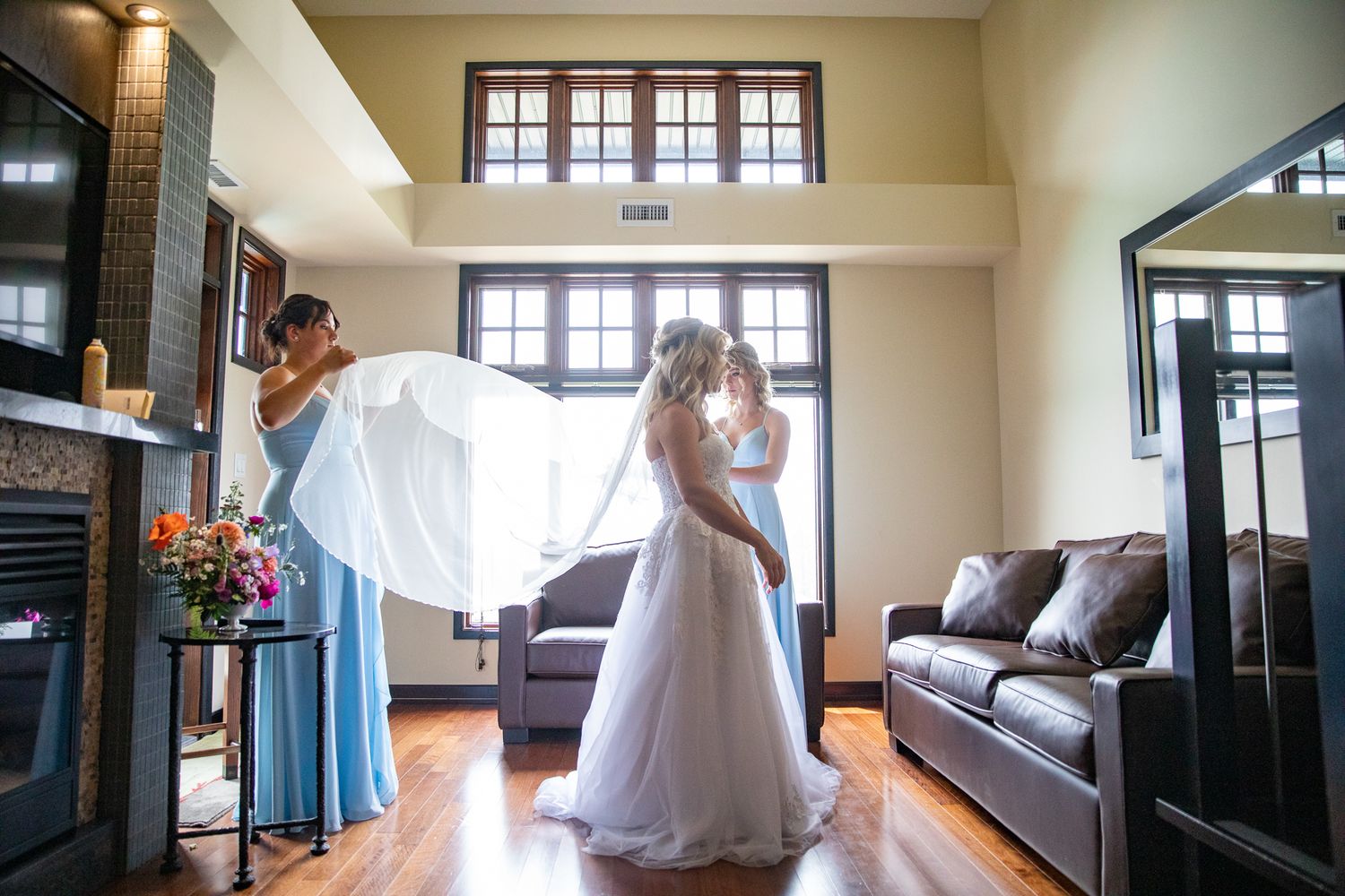Bride in white gown with bridesmaids in blue dresses preparing in elegant room with hardwood floors and large windows. Copperpoint resort,