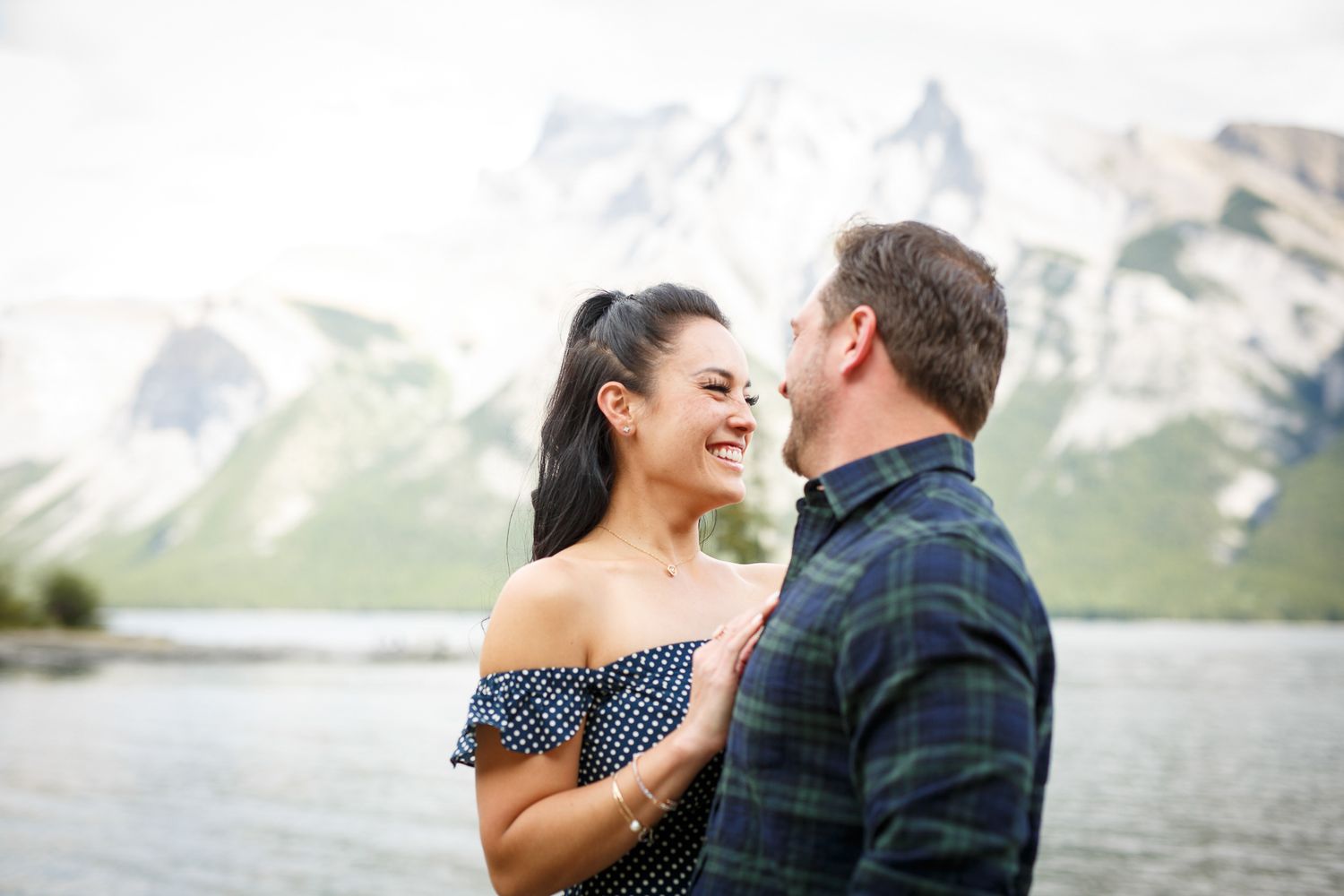 A smiling couple embracing outdoors near a body of water with mountains in the background at Lake Minnewanka in Banff.