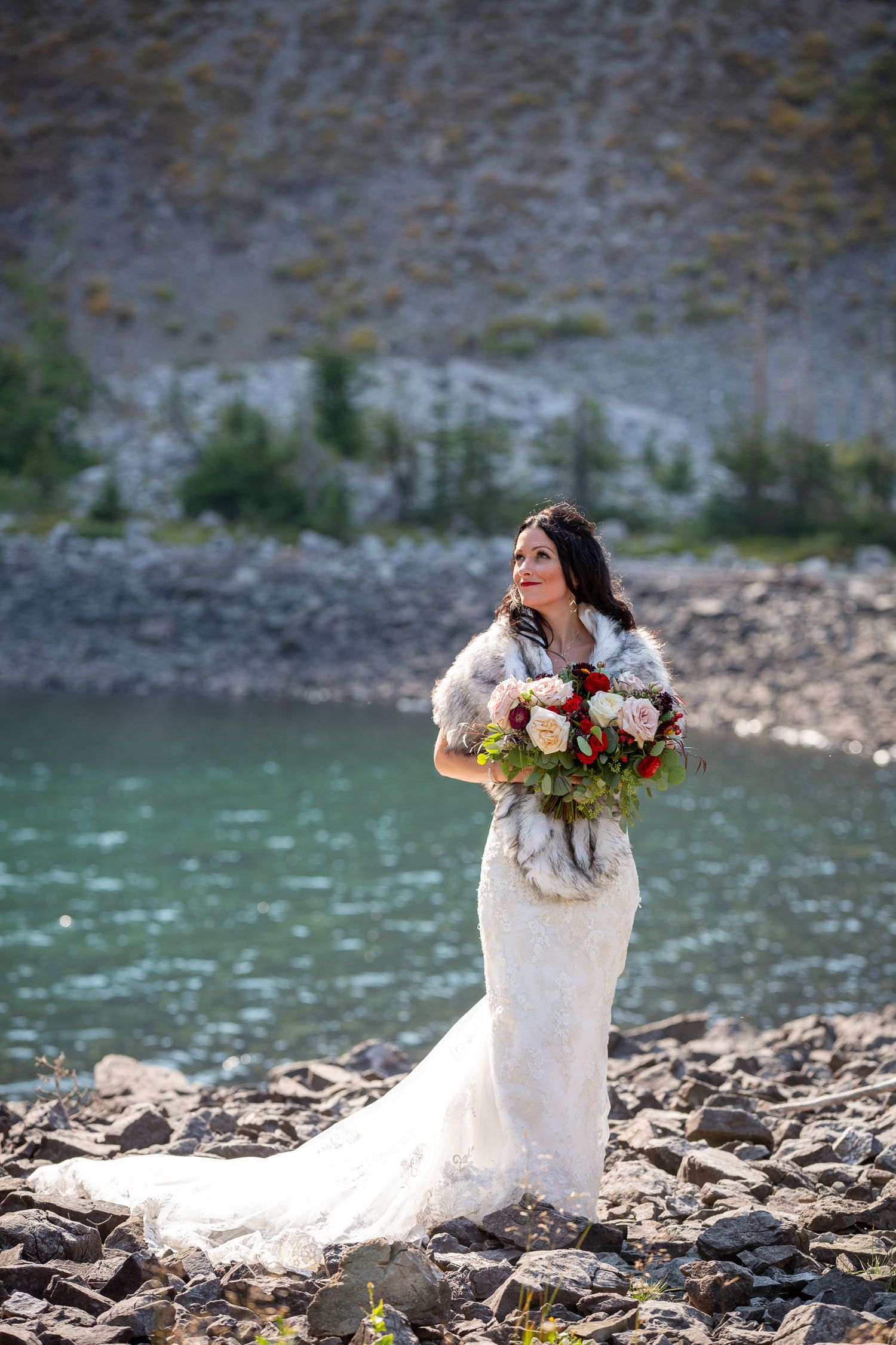 Bride in white lace gown with fur wrap holds bouquet by turquoise mountain lake on rocky shore in Kananaskis, Alberta.