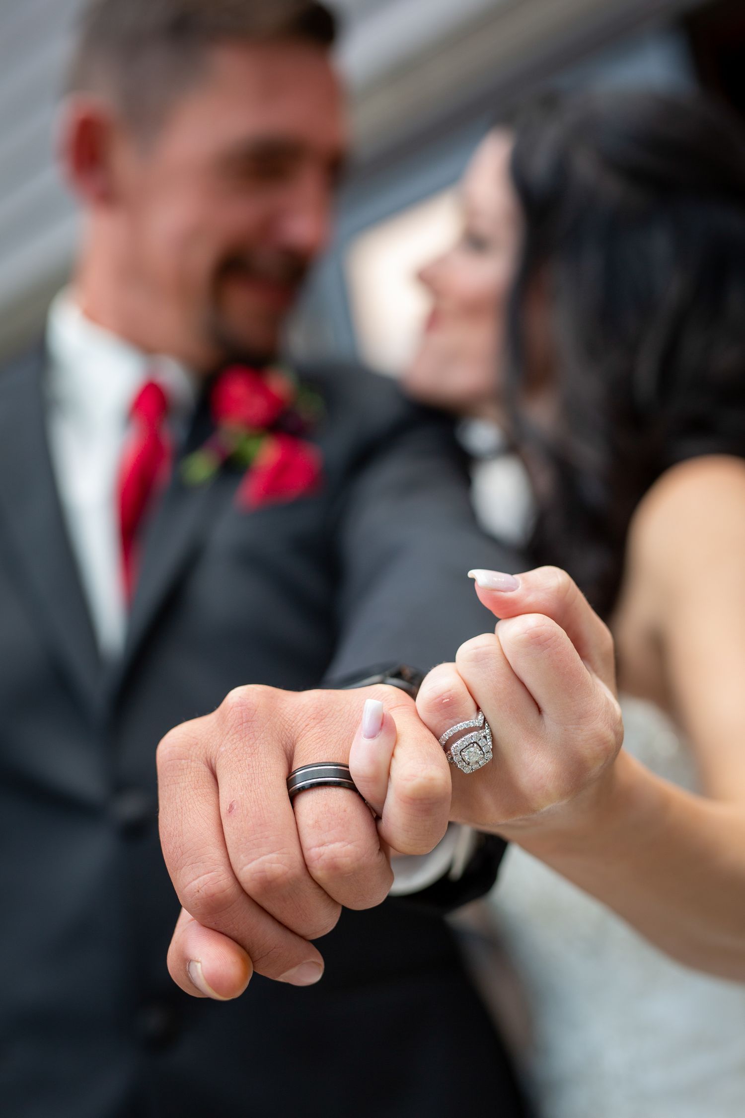 Newlyweds displaying wedding rings, groom in gray suit with red tie, bride with diamond ring, smiling couple in soft focus. Canmore, AB.