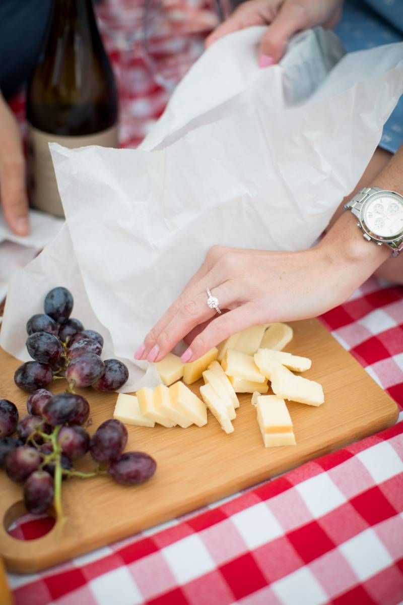 Hands unwrapping cheese on a wooden cutting board with red grapes and wine at a picnic at Fish Creek Park in Calgary.