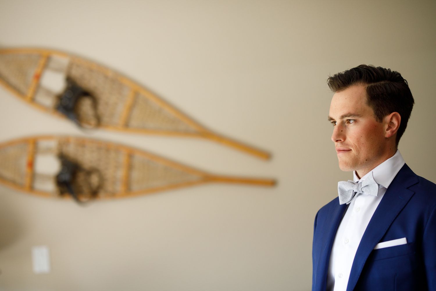 A groom wearing a navy blue suit and bow tie stands in front of a wooden wall with hanging decorations in Canmore.