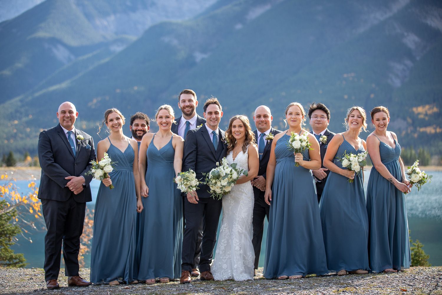Wedding party in dusty blue bridesmaid dresses and dark suits pose together against mountain backdrop at Quarry lake in Canmore, AB.