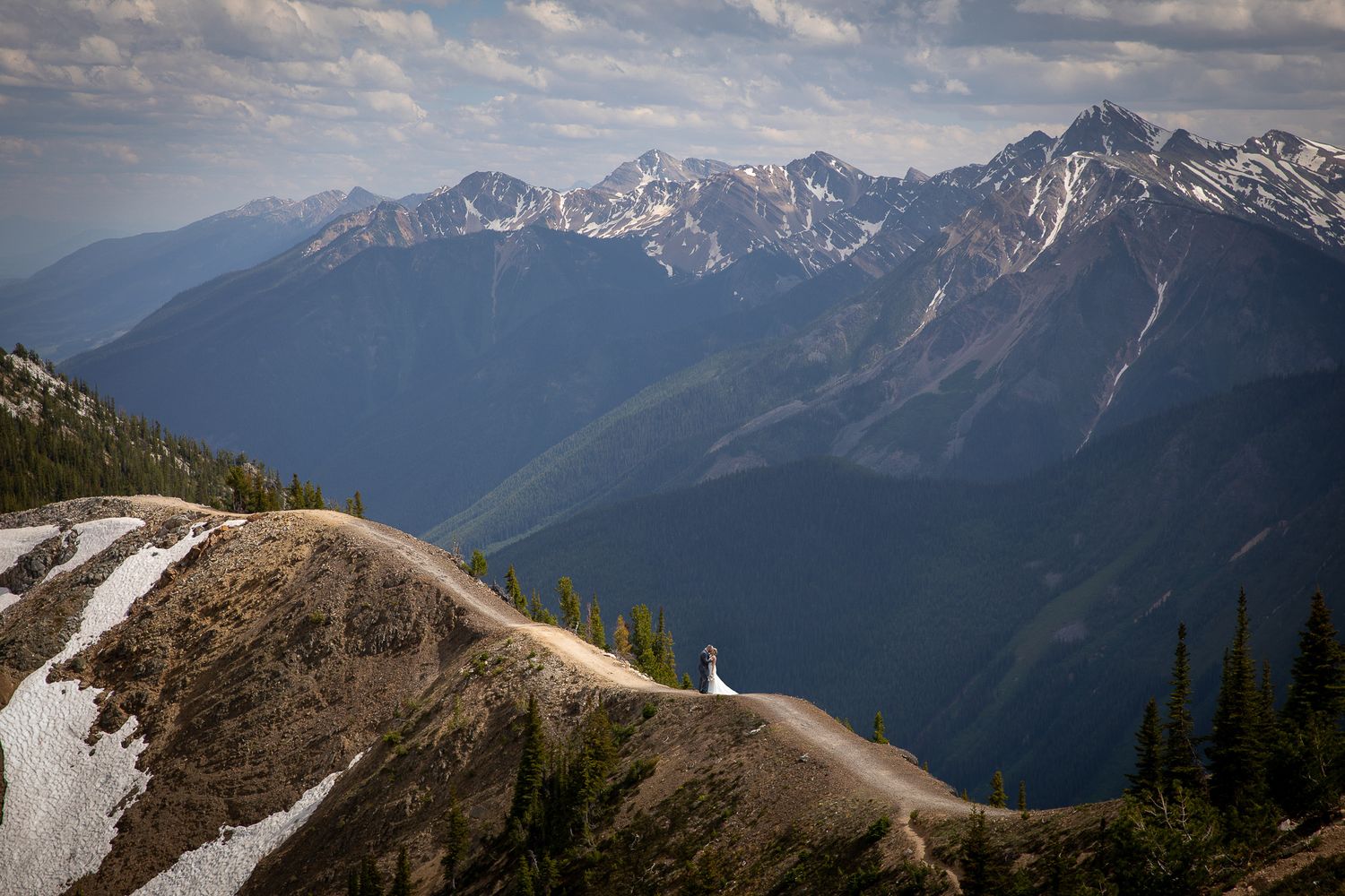 A wedding couple stands on a ridge trail overlooking majestic snow-capped peaks in the distance at Kicking Horse resort in Golden, BC.