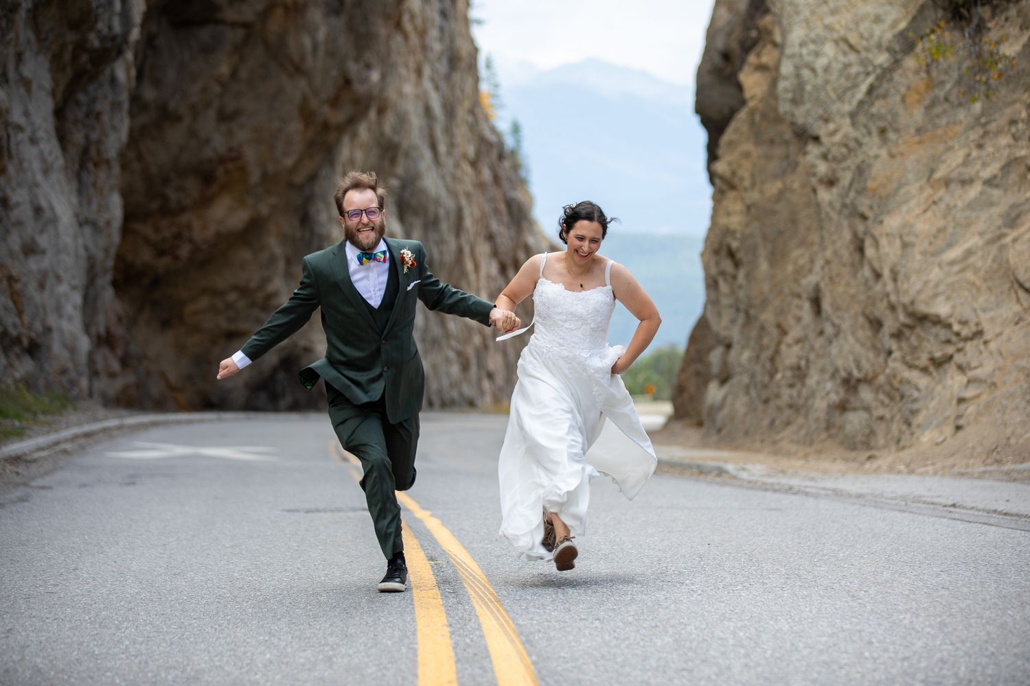 A couple in formal wedding attire runs playfully along a winding mountain road with steep cliffs at Sinclair canyon in Radium, BC.
