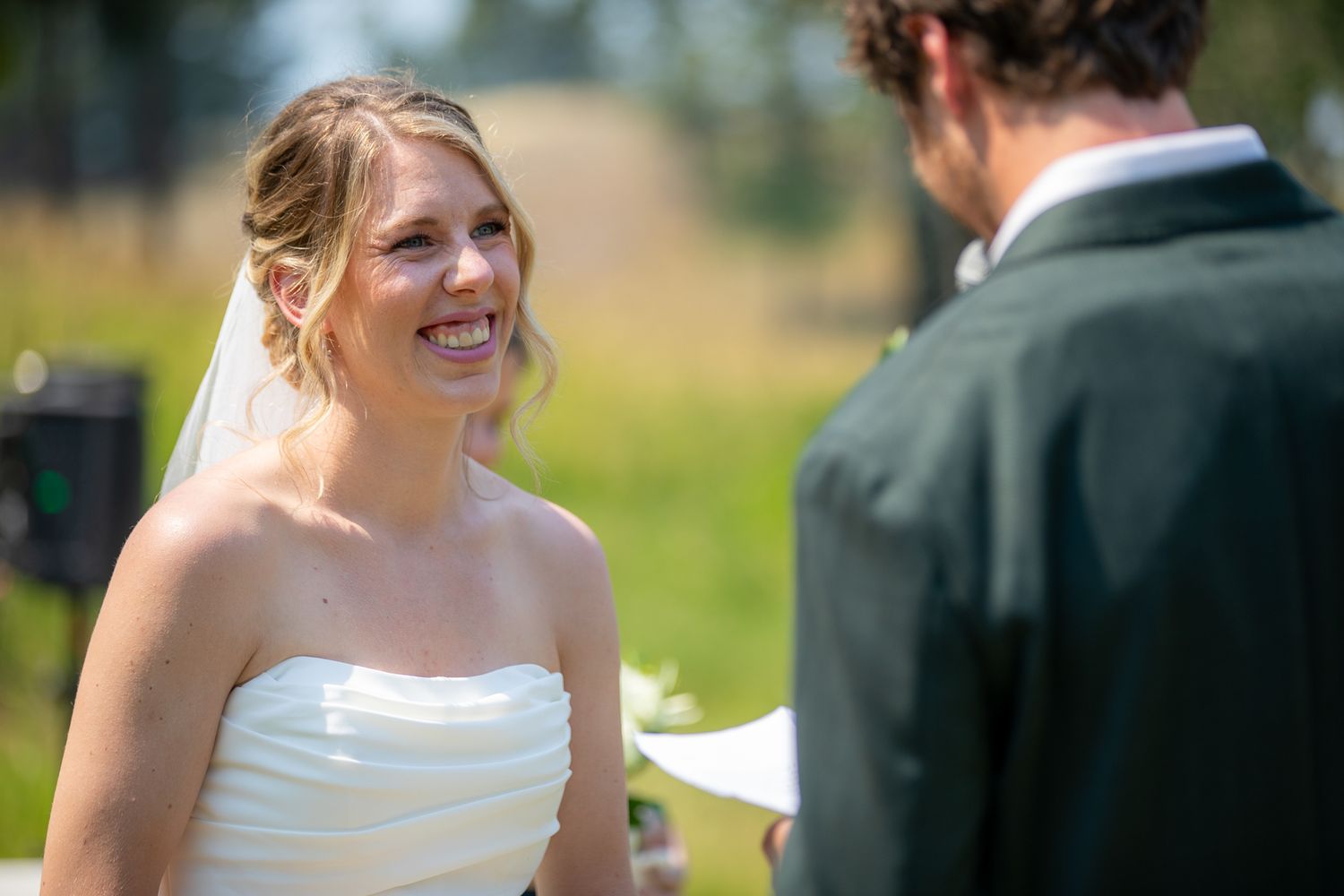 Smiling bride in white strapless gown and veil listening to groom read vows at outdoor wedding ceremony at Swansea Ranch in Invermere, BC