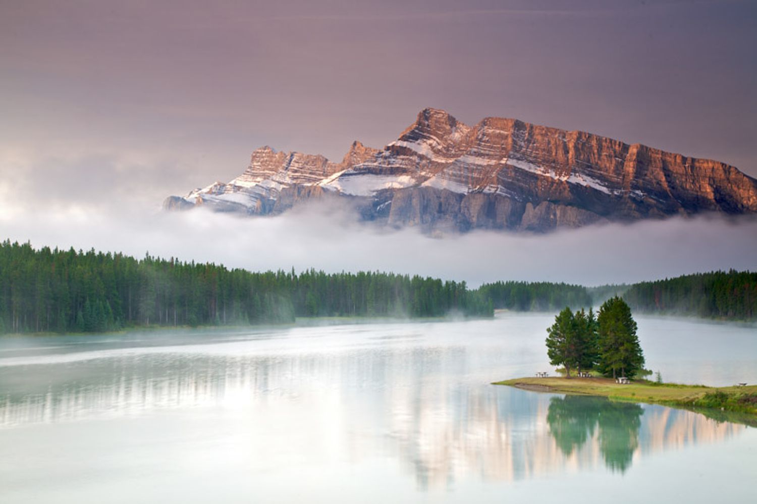 Scenic mountain Two Jack lake panorama with misty clouds reflecting in calm waters during sunrise at Banff National Park.